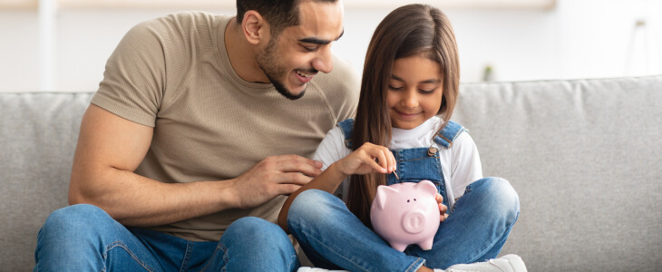 Father sitting with daughter who is putting a coin in a piggy bank