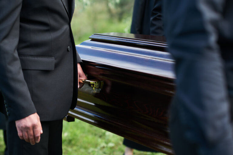 Focus on man in black suit holding by handle of wooden coffin