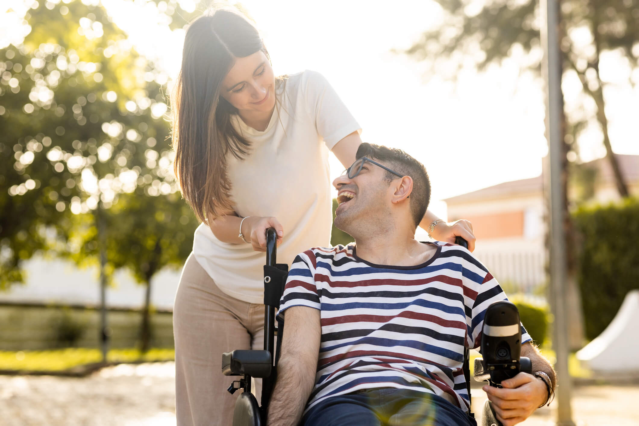 An adult man with a disability sits in an automatic wheelchair with a young woman in a park at sunset.The woman and the man look at each other happily.