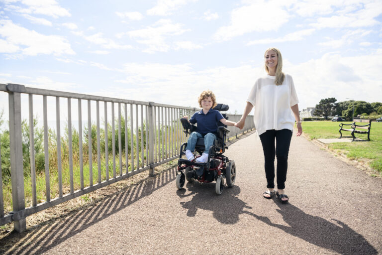 Portrait of 6 year old disabled boy with muscular dystrophy, operating electric wheelchair while holding hands with his mid adult mother in the sunlight