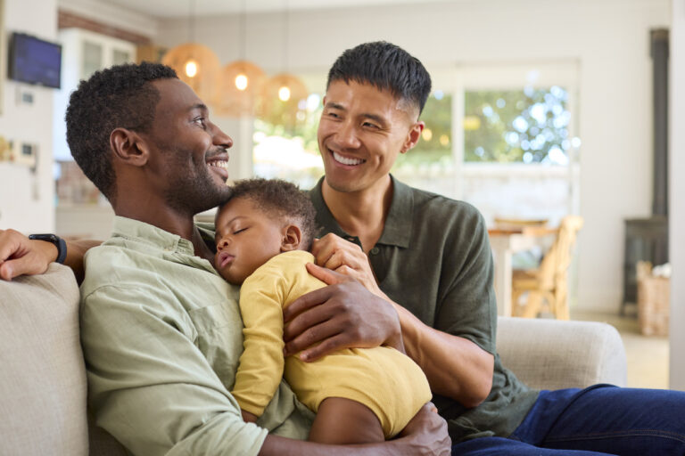 Same Sex Male Couple Cuddling Sleeping Baby Daughter On Sofa