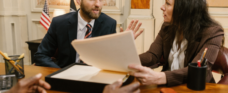 Two people in suits discuss documents at a wooden conference table.