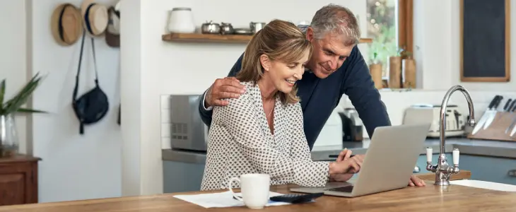 Middle aged couple sitting in kitchen looking at laptop.