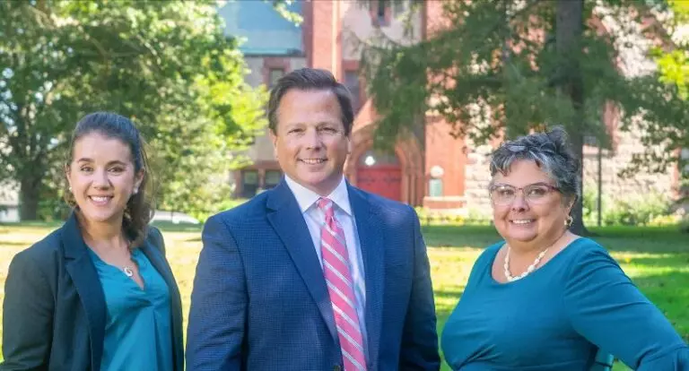 A professional photo of two women and a man in business attire, smiling at the camera outdoors.