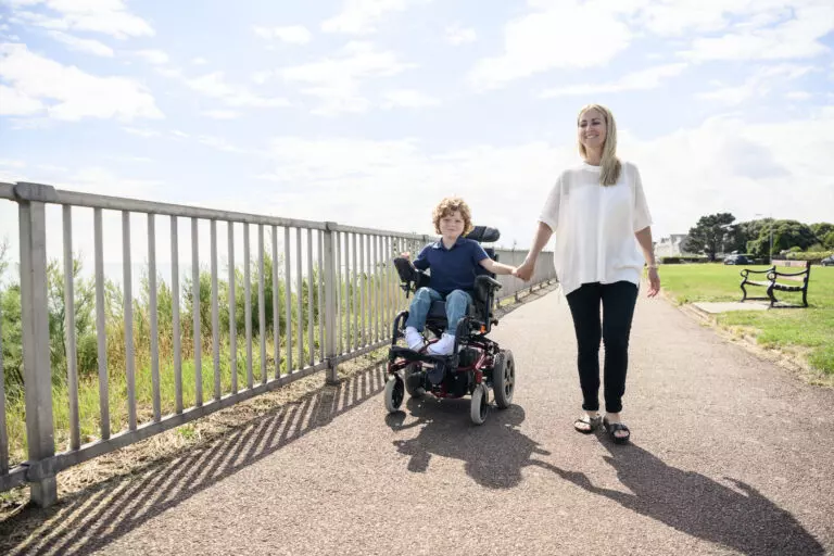 Portrait of 6 year old disabled boy with muscular dystrophy, operating electric wheelchair while holding hands with his mid adult mother in the sunlight