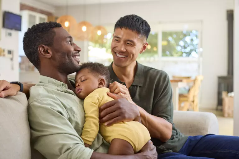 Same Sex Male Couple Cuddling Sleeping Baby Daughter On Sofa